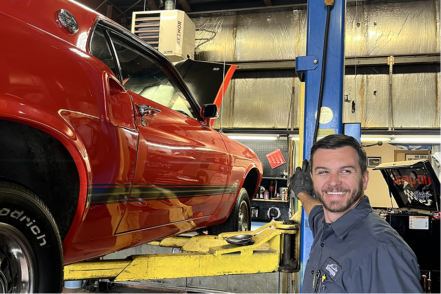 Man in mechanic shop next to a red car on a lift; smiling and pointing. | Acadia Auto Service