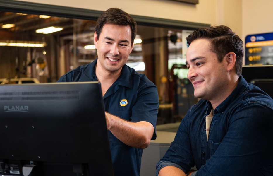 Two men smiling at a computer screen in an auto repair shop. One wears a NAPA uniform. | Acadia Auto Service