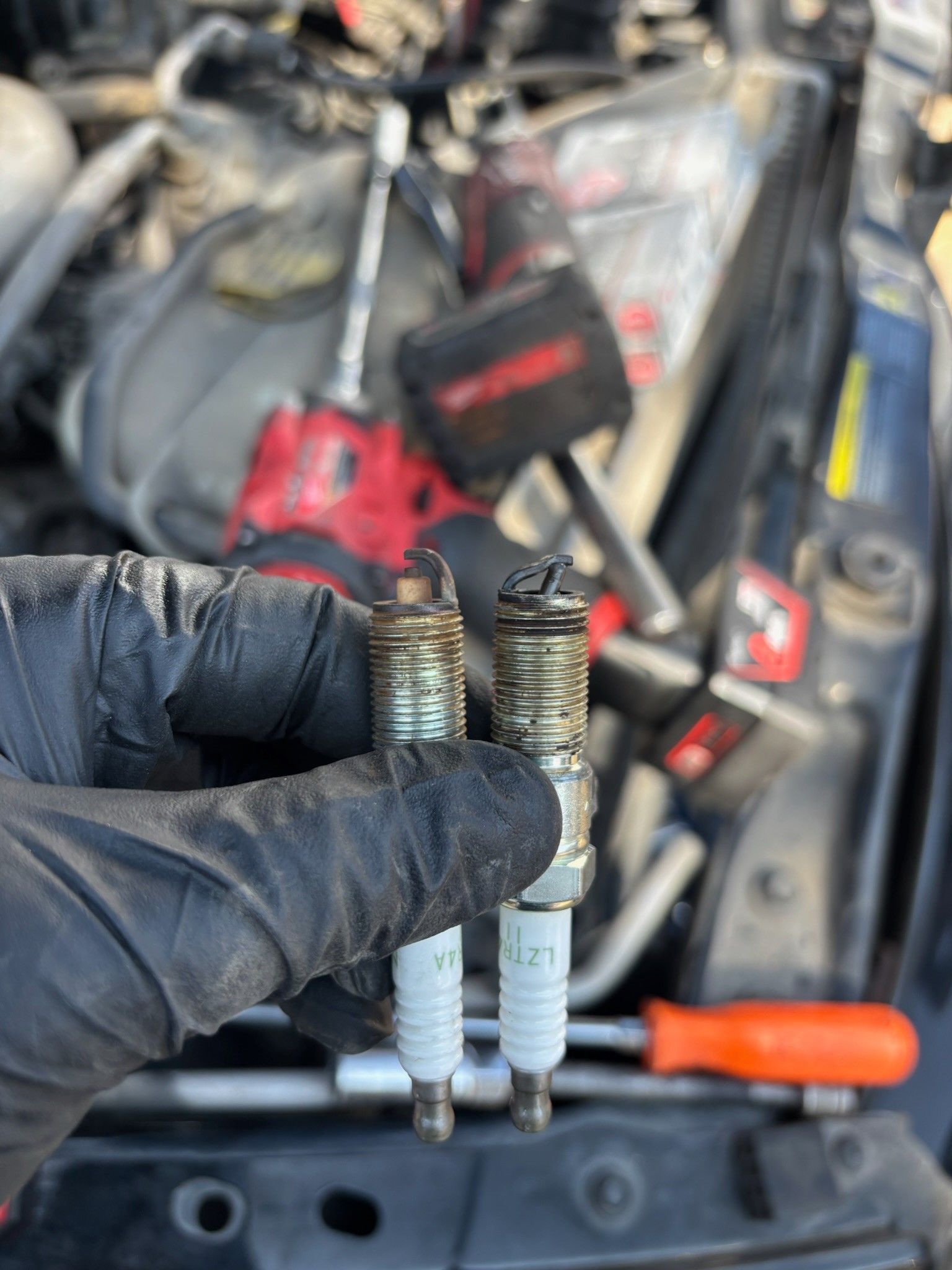 Gloved hand holding two worn spark plugs in a car engine bay, with tools blurred in the background