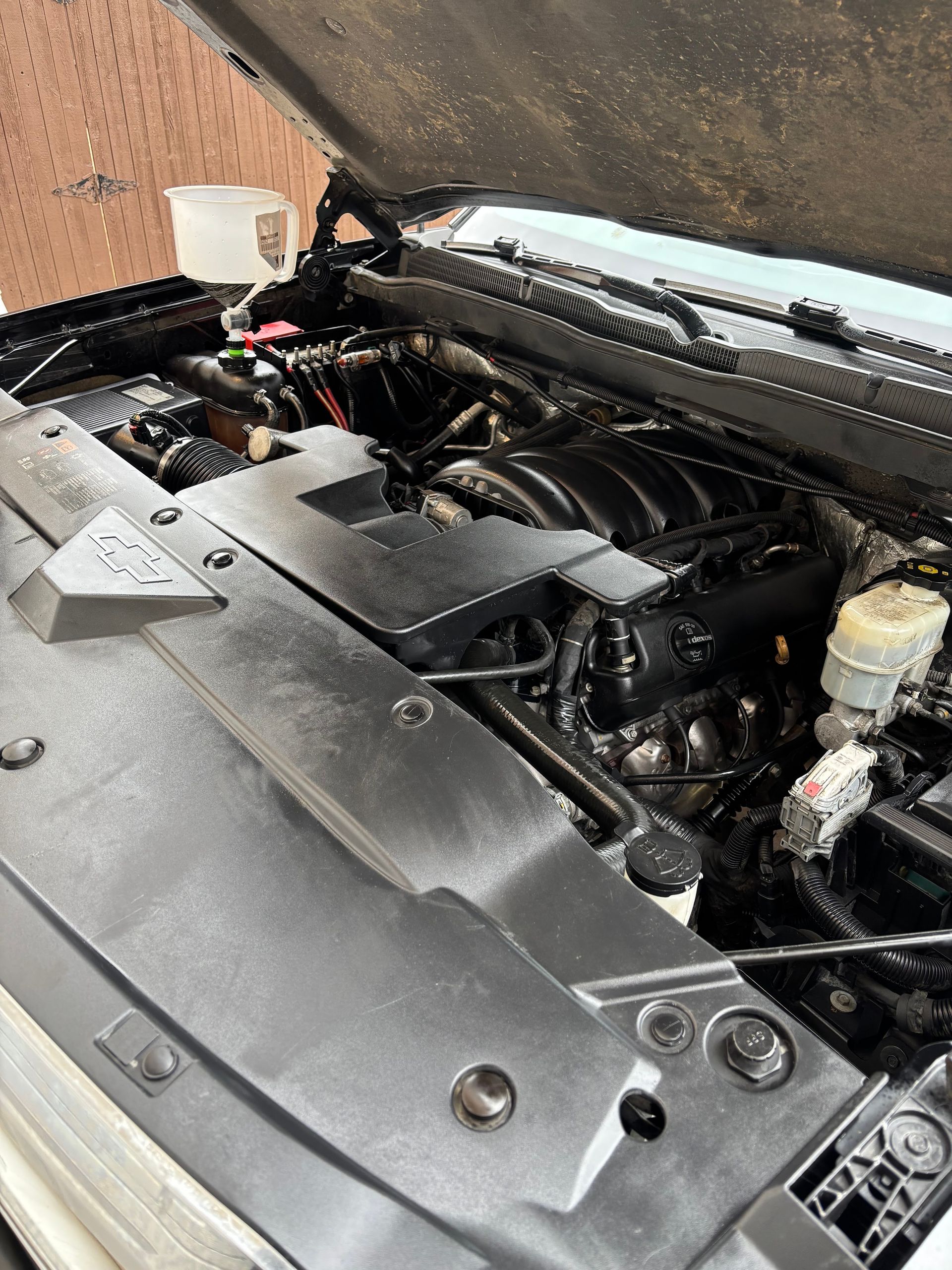 Car engine bay with the hood open, showing black hoses, silver covers, and fluid reservoirs.