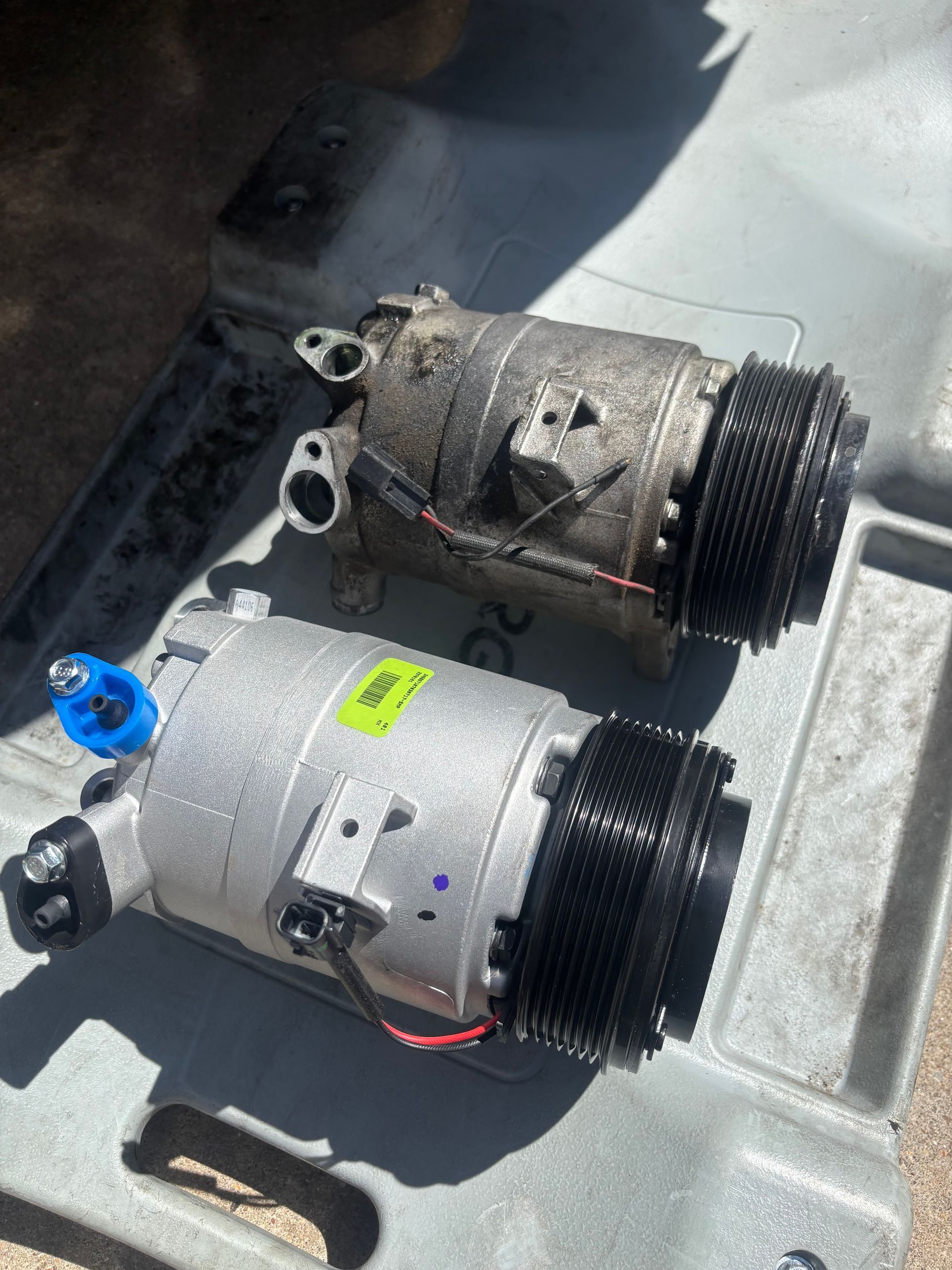 Two used gray automotive compressors lying in a truck bed, one with a blue cap and one weathered with rust.