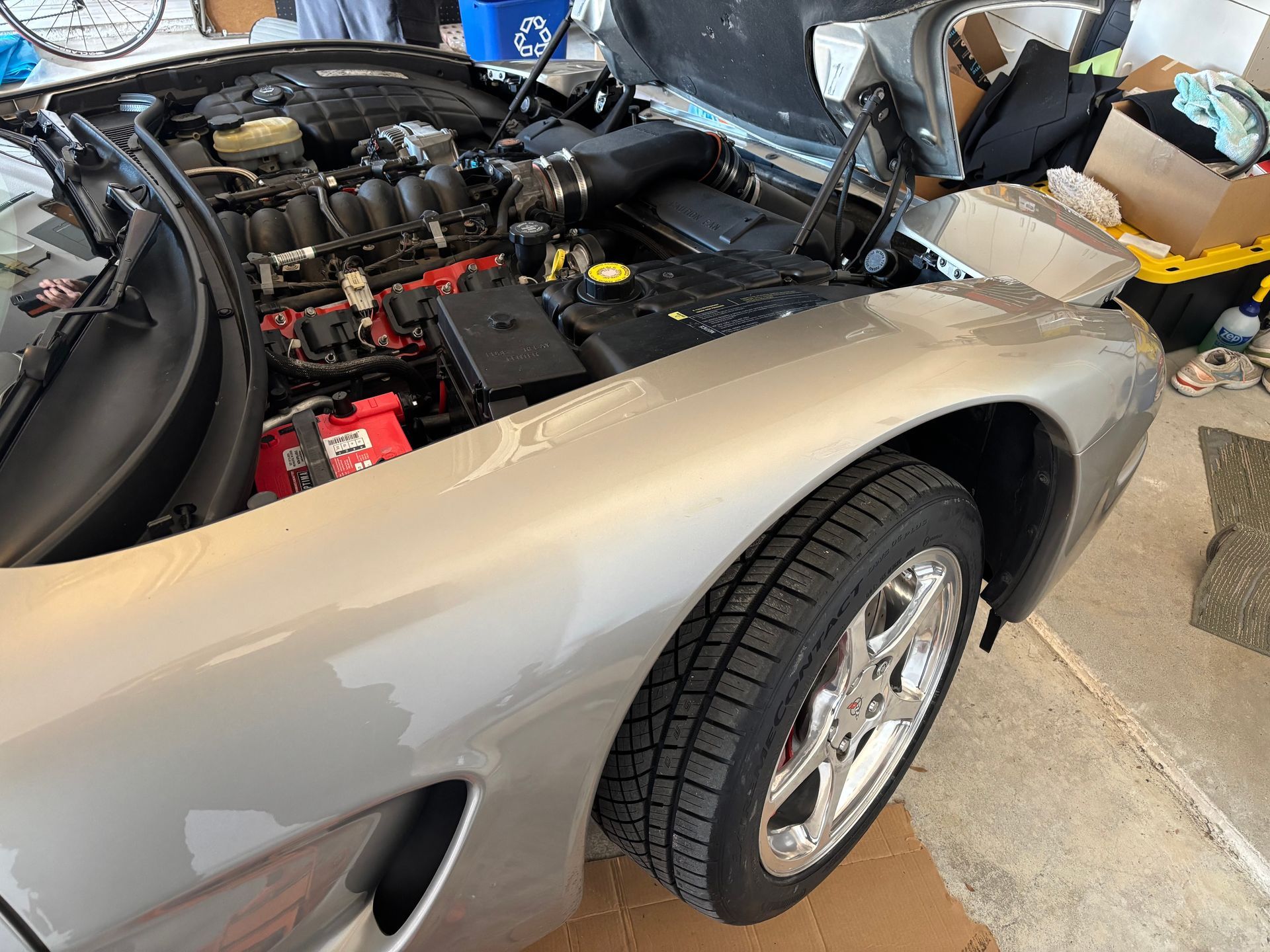 Silver sports car with open hood in a garage, showing the engine and front wheel.