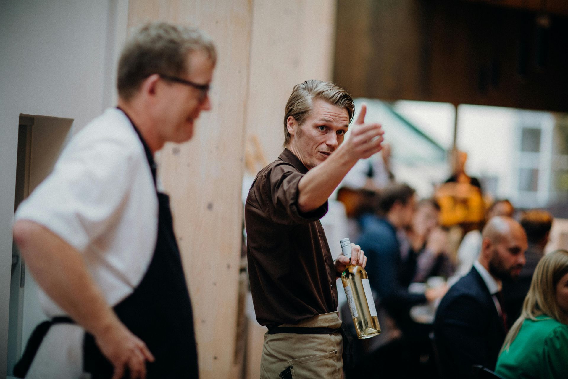 Twee mannen met schorten om, waarvan er één een duim omhoog steekt terwijl hij een fles vasthoudt.