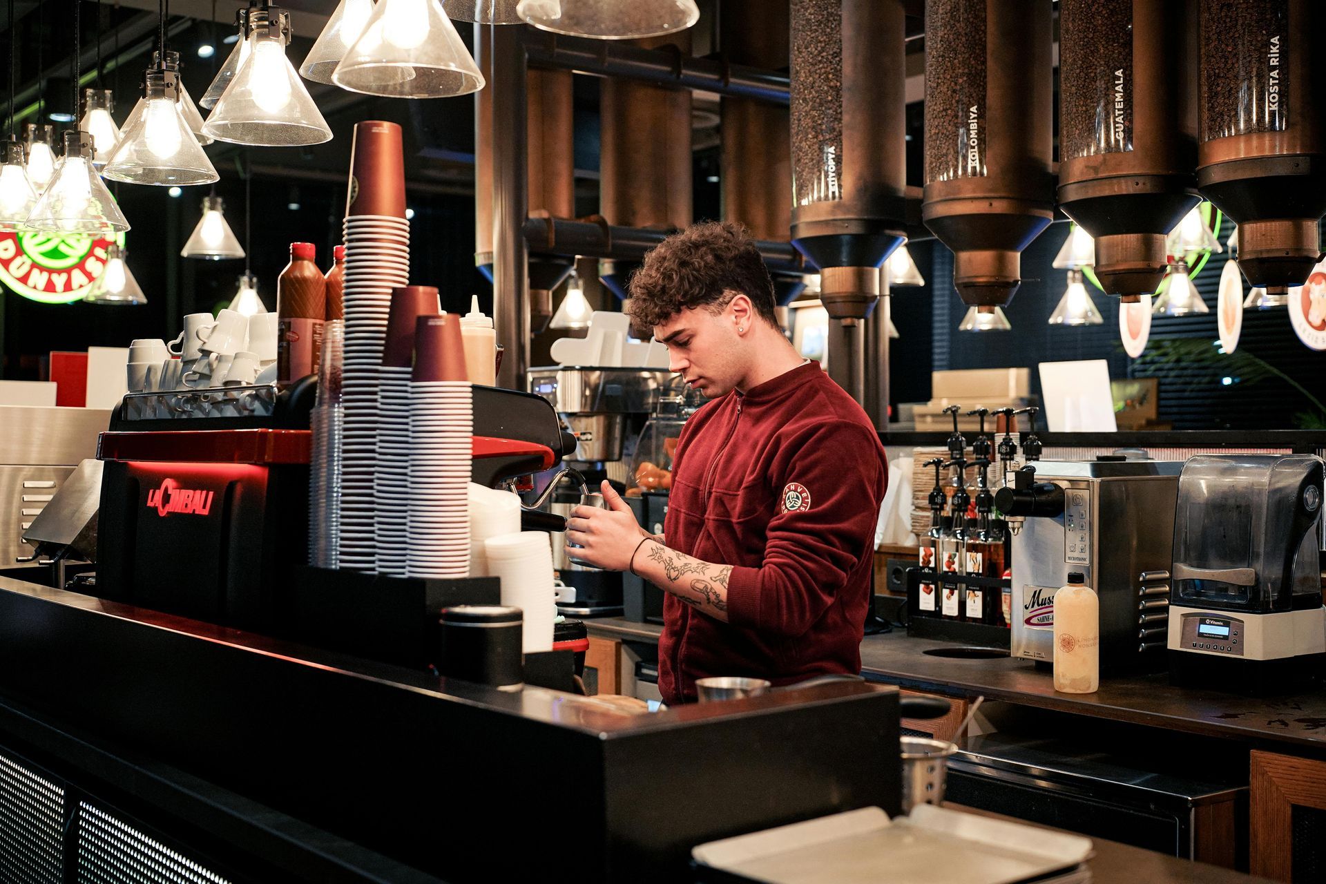 Een barman in een rode trui maakt een drankje klaar achter een schemerig verlichte bar met hangende lampen.