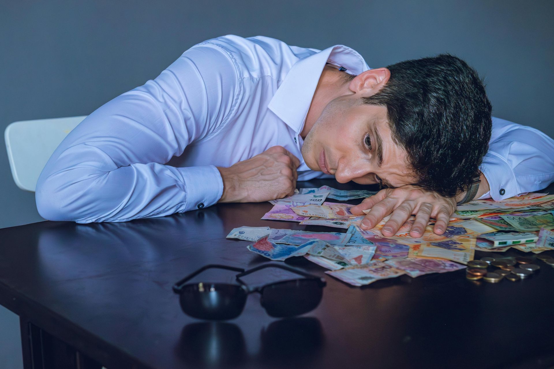 Een persoon in een wit overhemd laat zijn hoofd rusten op een tafel vol verspreid geld, met een verm