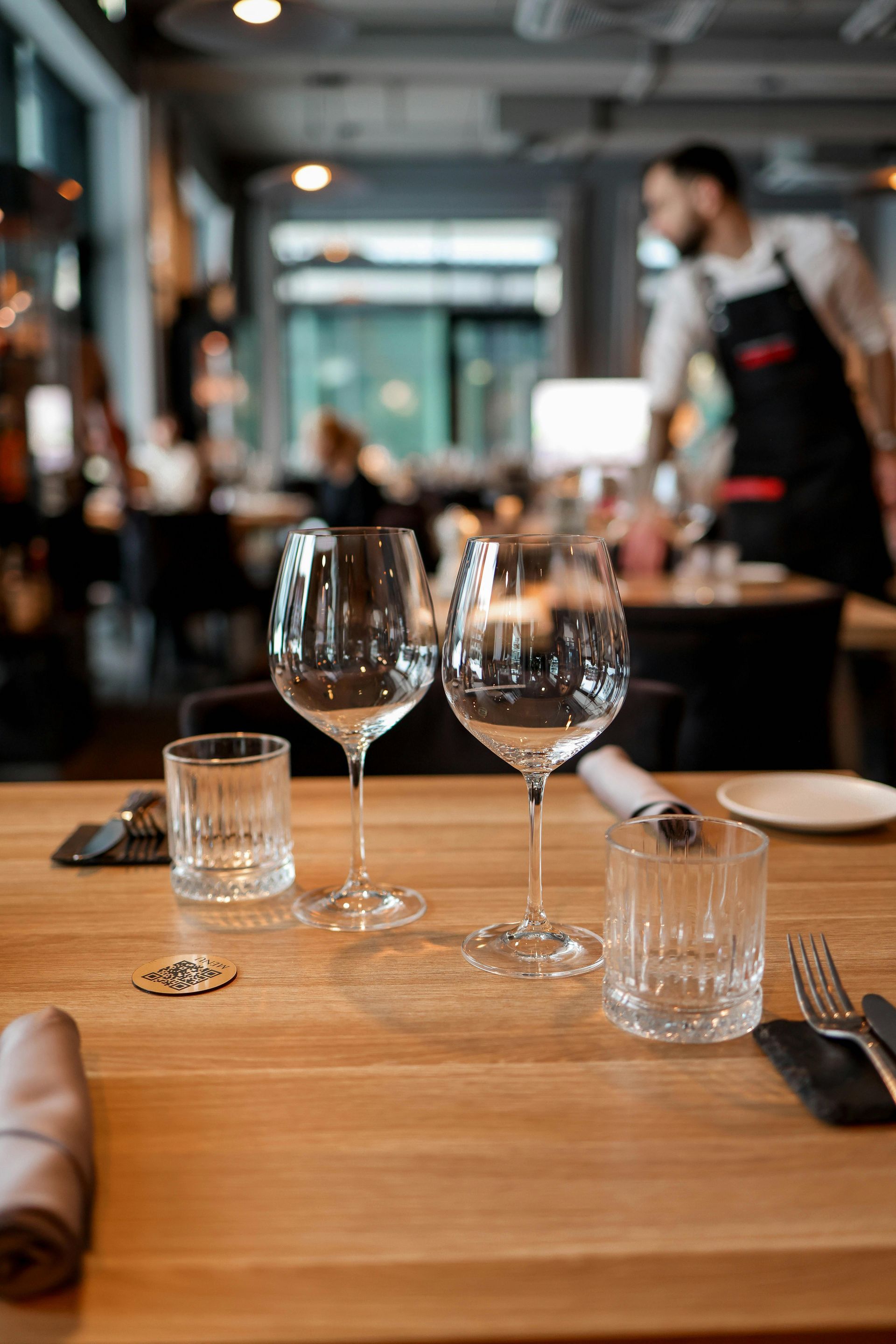 Een gedekte eettafel met wijnglazen en waterglazen in een restaurant; een ober is wazig op de achte