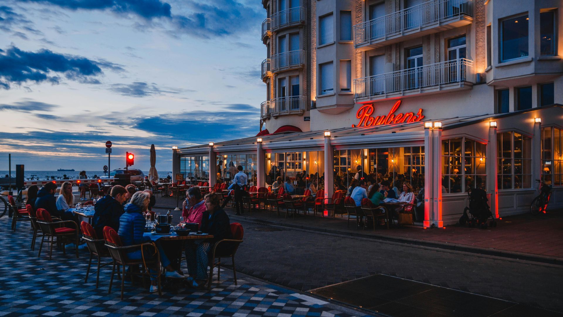 Een terrasje aan het water bij schemering, met gasten onder warme verlichting en een oplichtend restaurantbord.