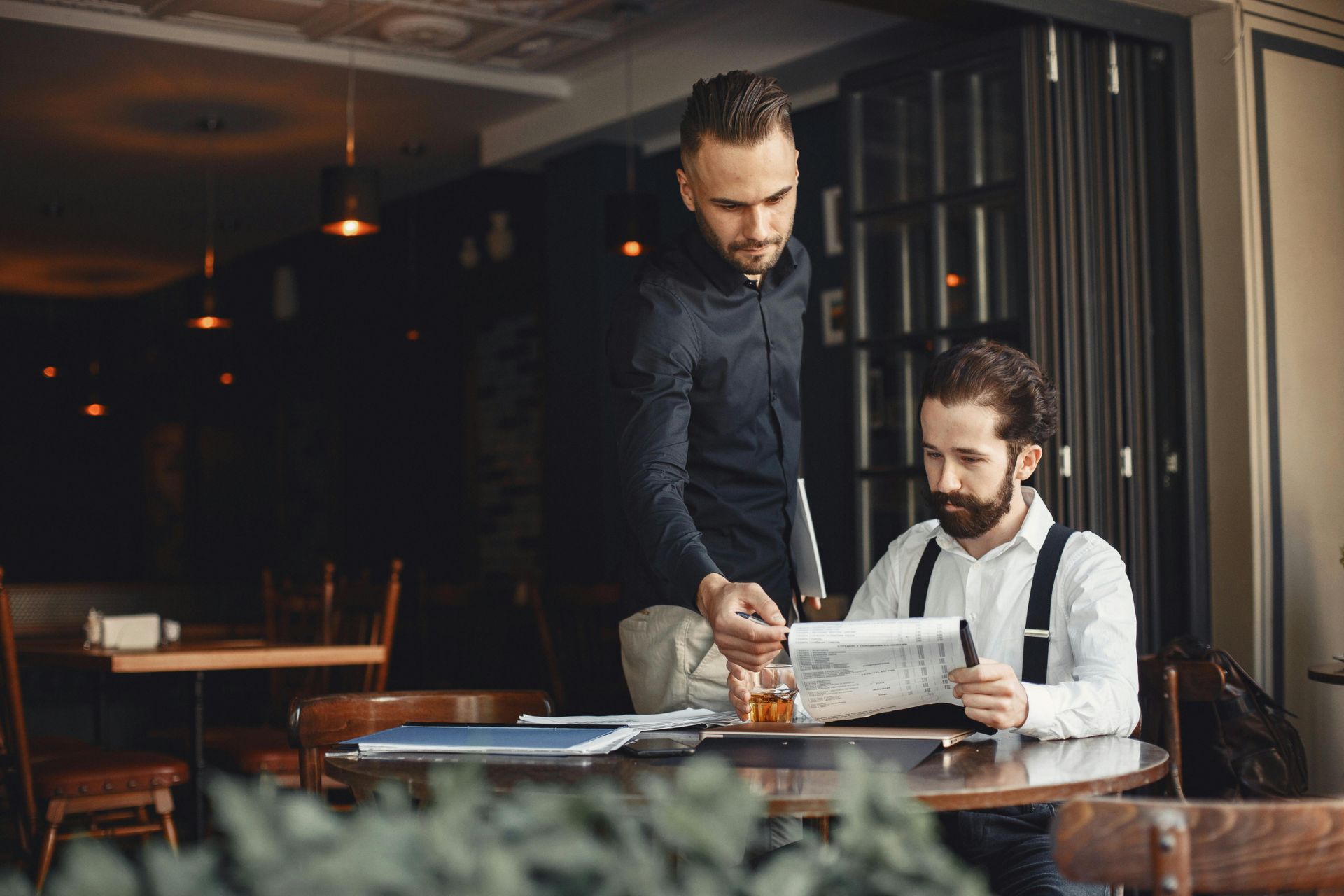 Twee mannen bekijken documenten aan een restauranttafel; de ene staat en wijst, de andere leest
