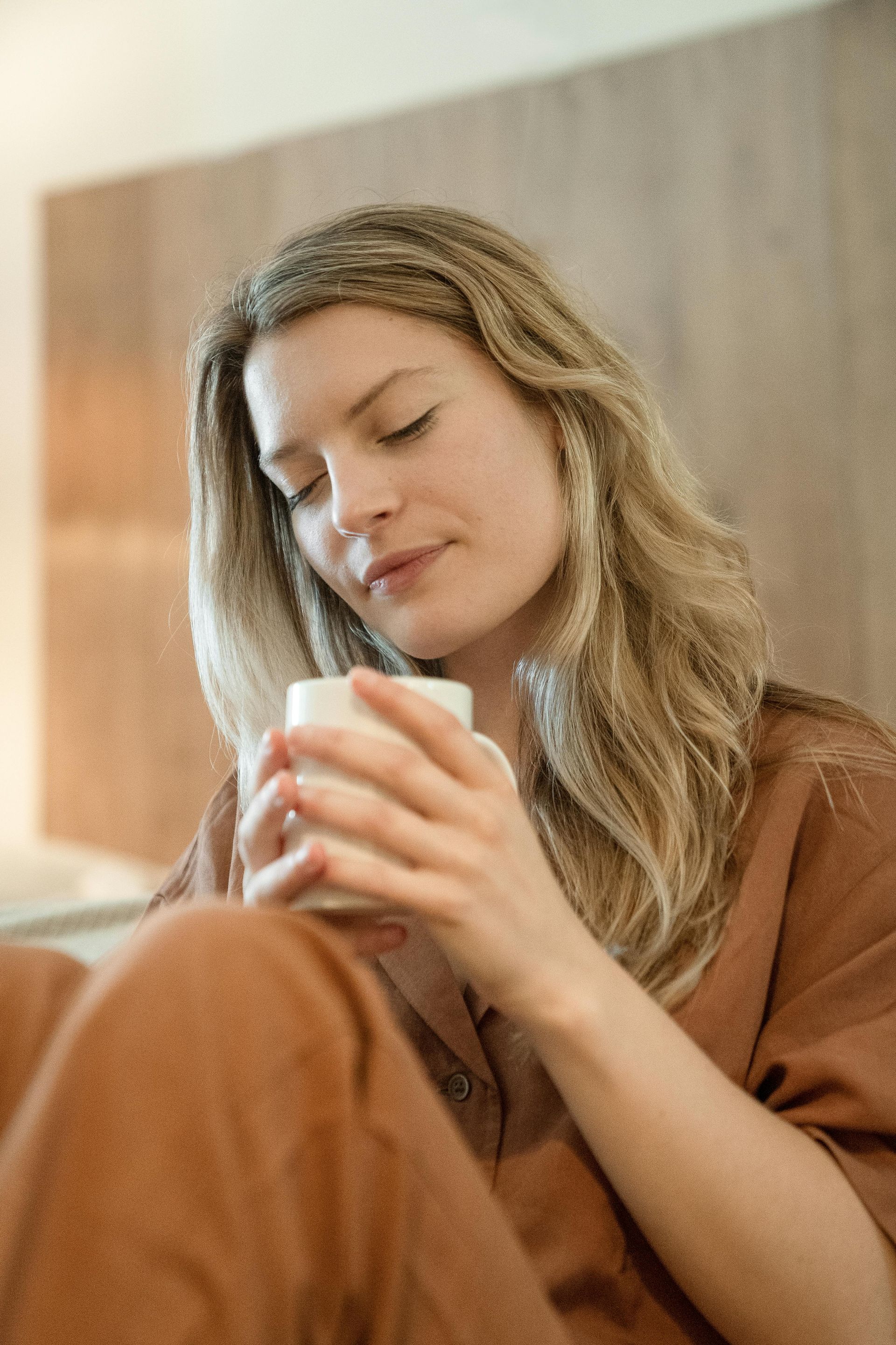 Een persoon met lang golvend haar, gekleed in een bruin shirt, houdt met beide handen een witte mok vast, met gesloten ogen en een kalme uitdrukking.