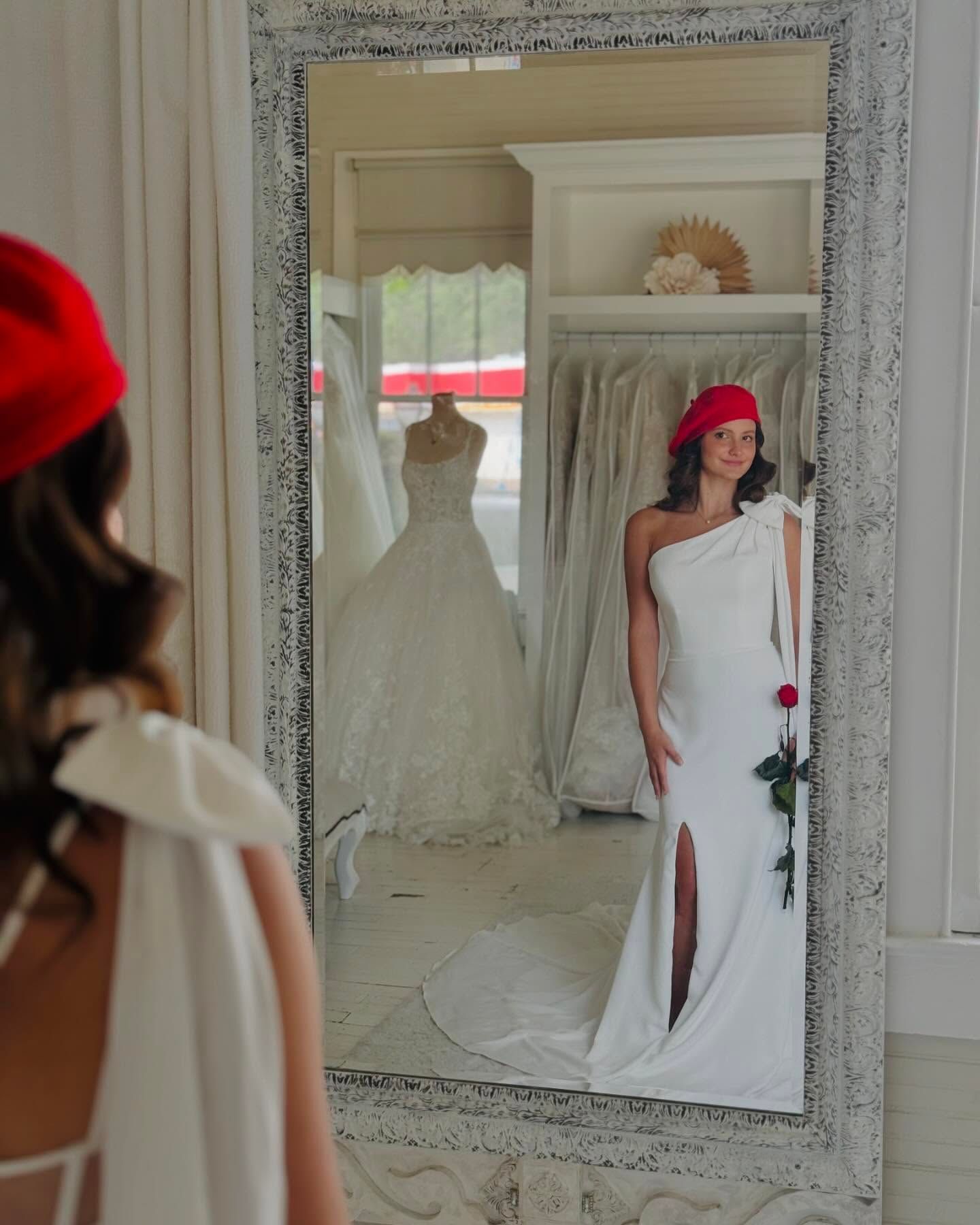 Woman in a white gown and red beret, posing in a mirror inside a bridal shop.