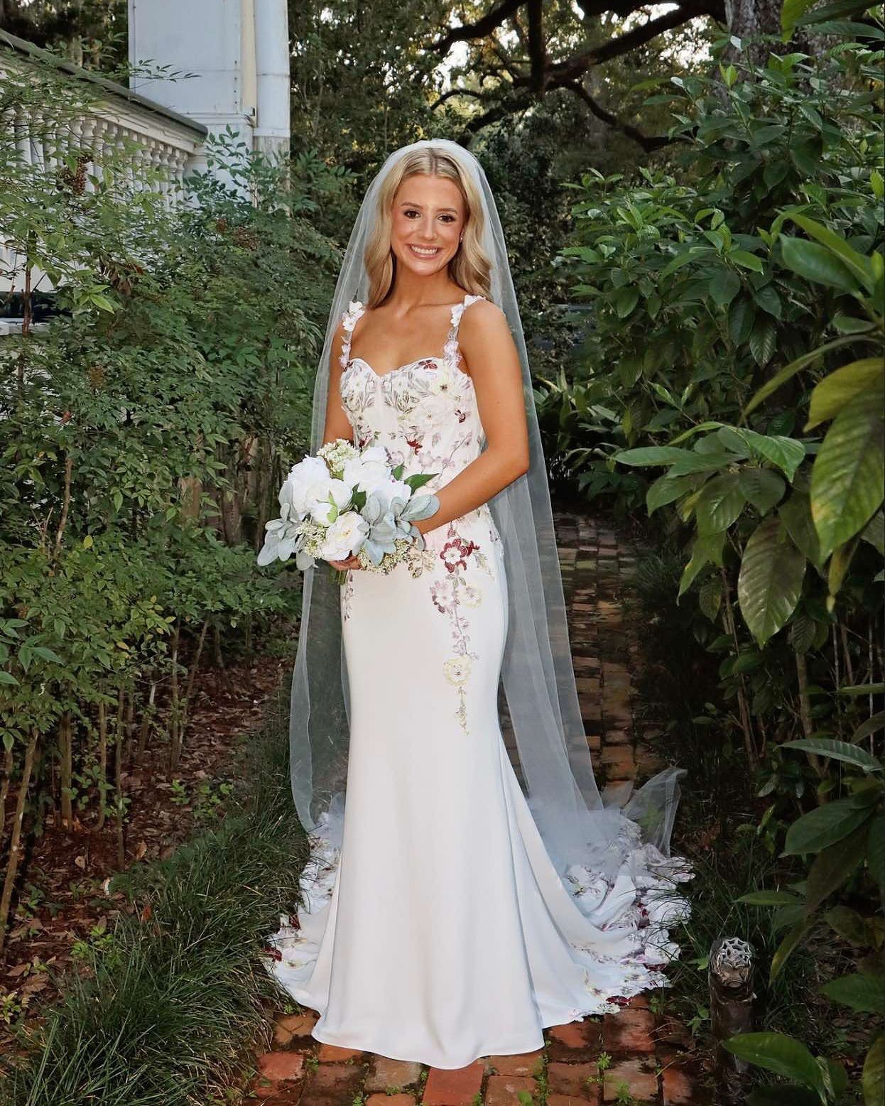 Blonde bride in white gown with floral bodice, holding bouquet, long veil, standing outdoors.