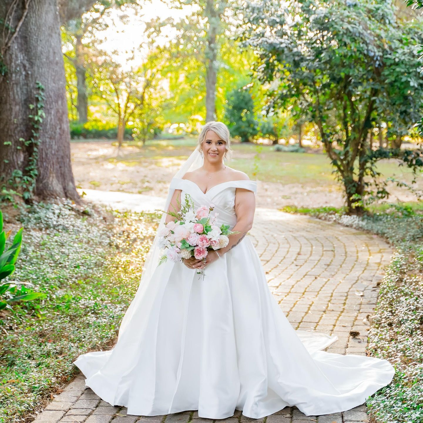 Bride in white dress, holding bouquet, smiling in a garden with stone pathway.