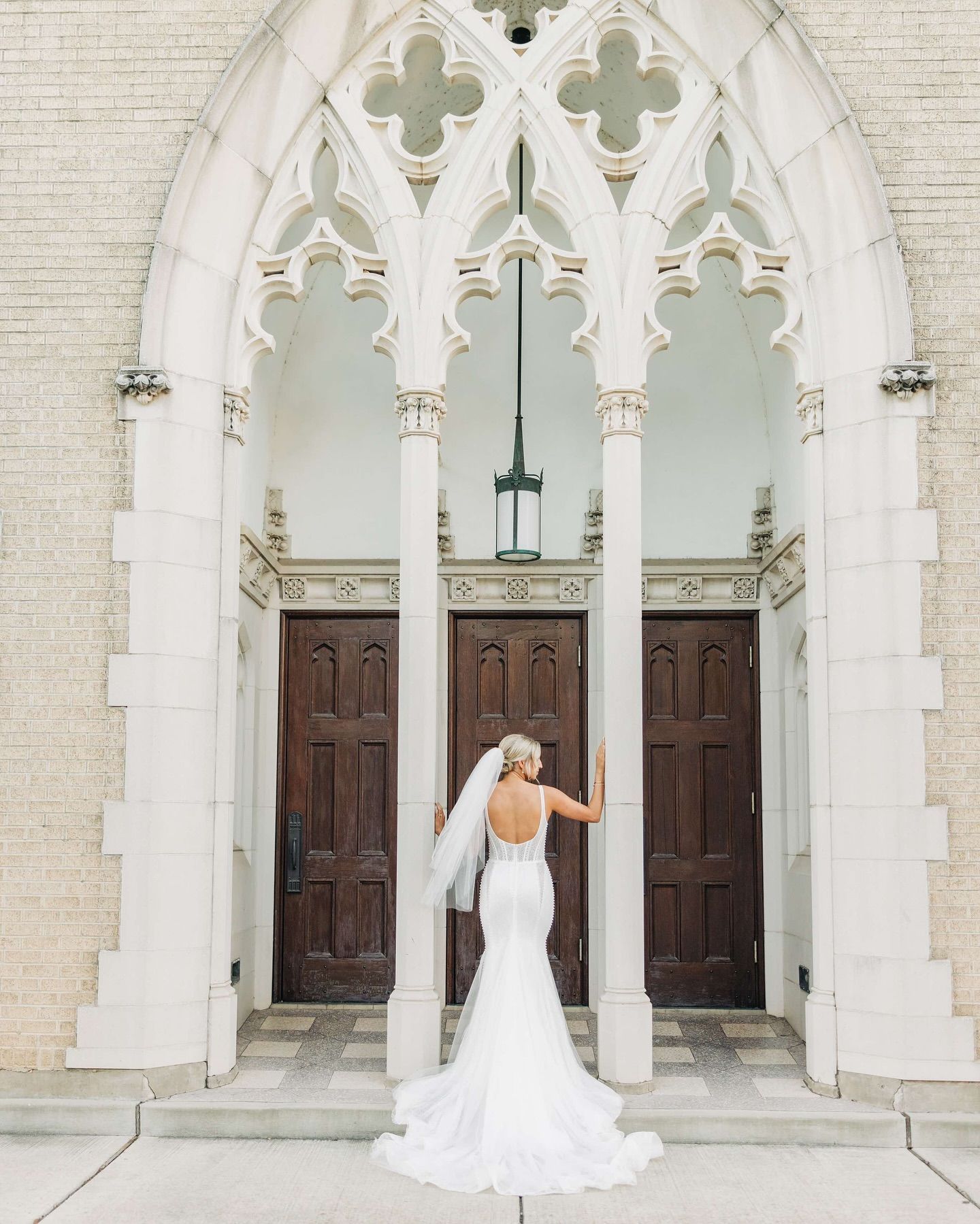 Bride in a wedding dress stands under a church archway, arms outstretched.
