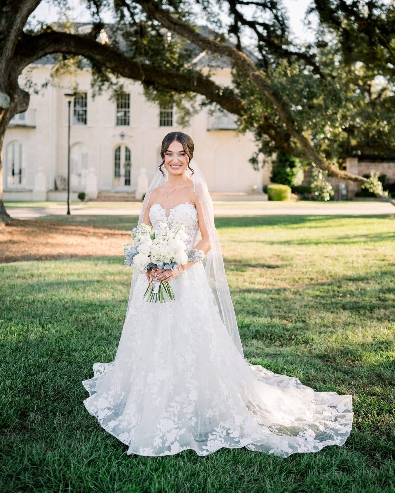 Bride in white lace dress, holding bouquet, smiles in front of a white building and green lawn.