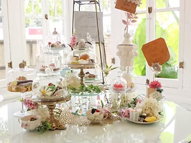 Pastries on tiered glass stands, floral arrangements on a white table, near a window.