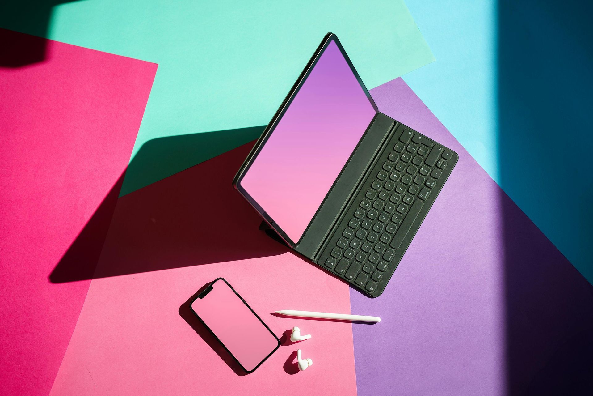 A laptop , cell phone , ear buds , and pen are on a colorful surface.