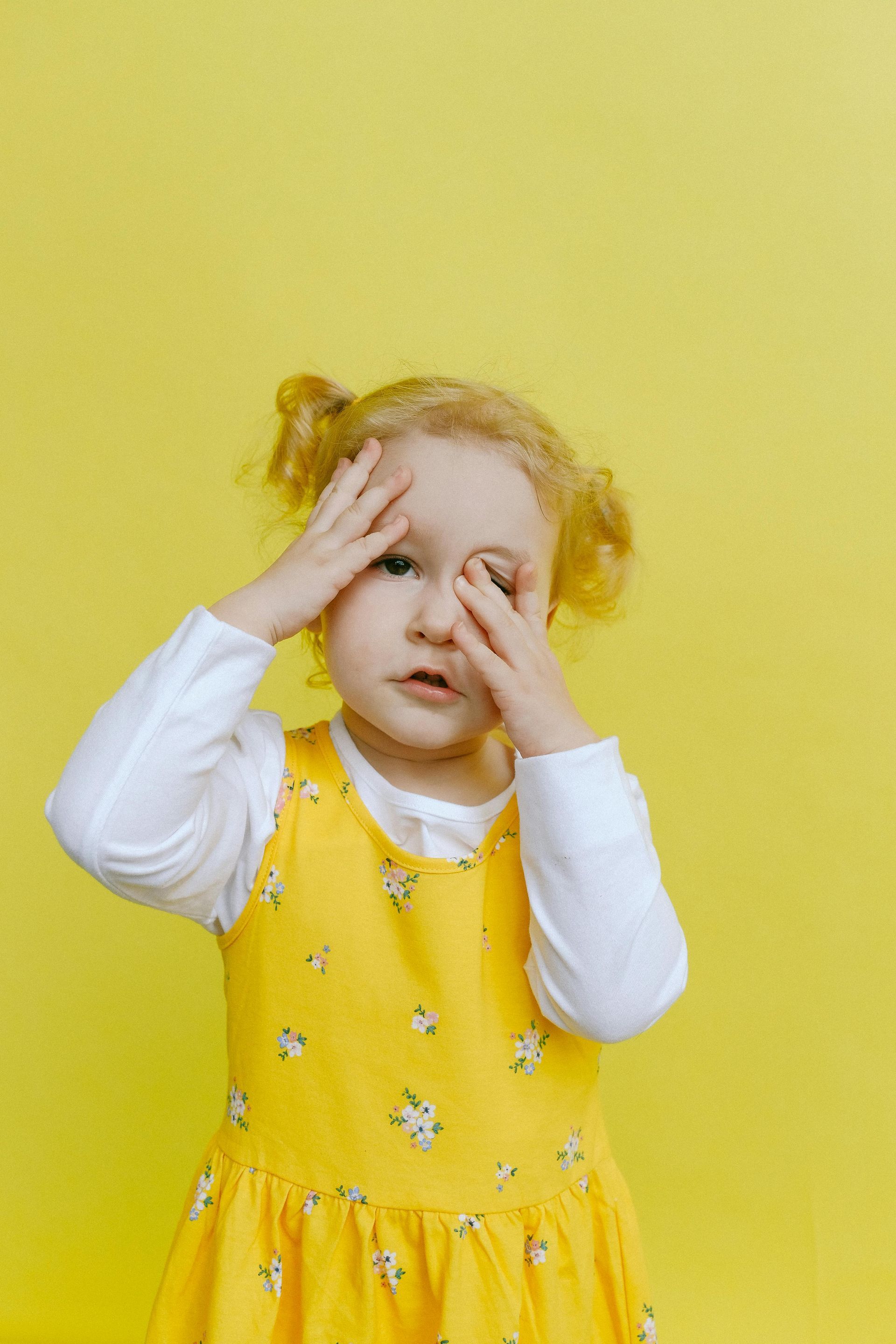 A little girl in a yellow dress is covering her eyes with her hands.