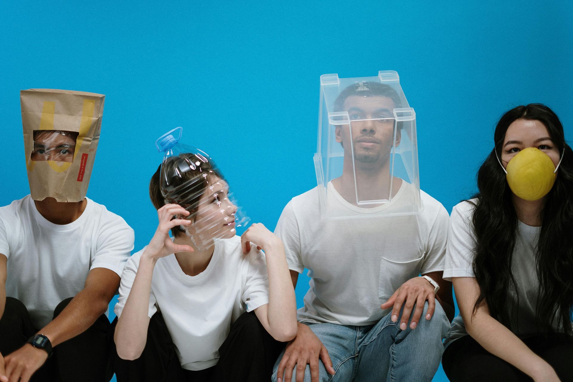 Four people sit, each wearing a makeshift mask: paper bag, plastic bottle, clear shield, and a yellow mask, against a blue background.