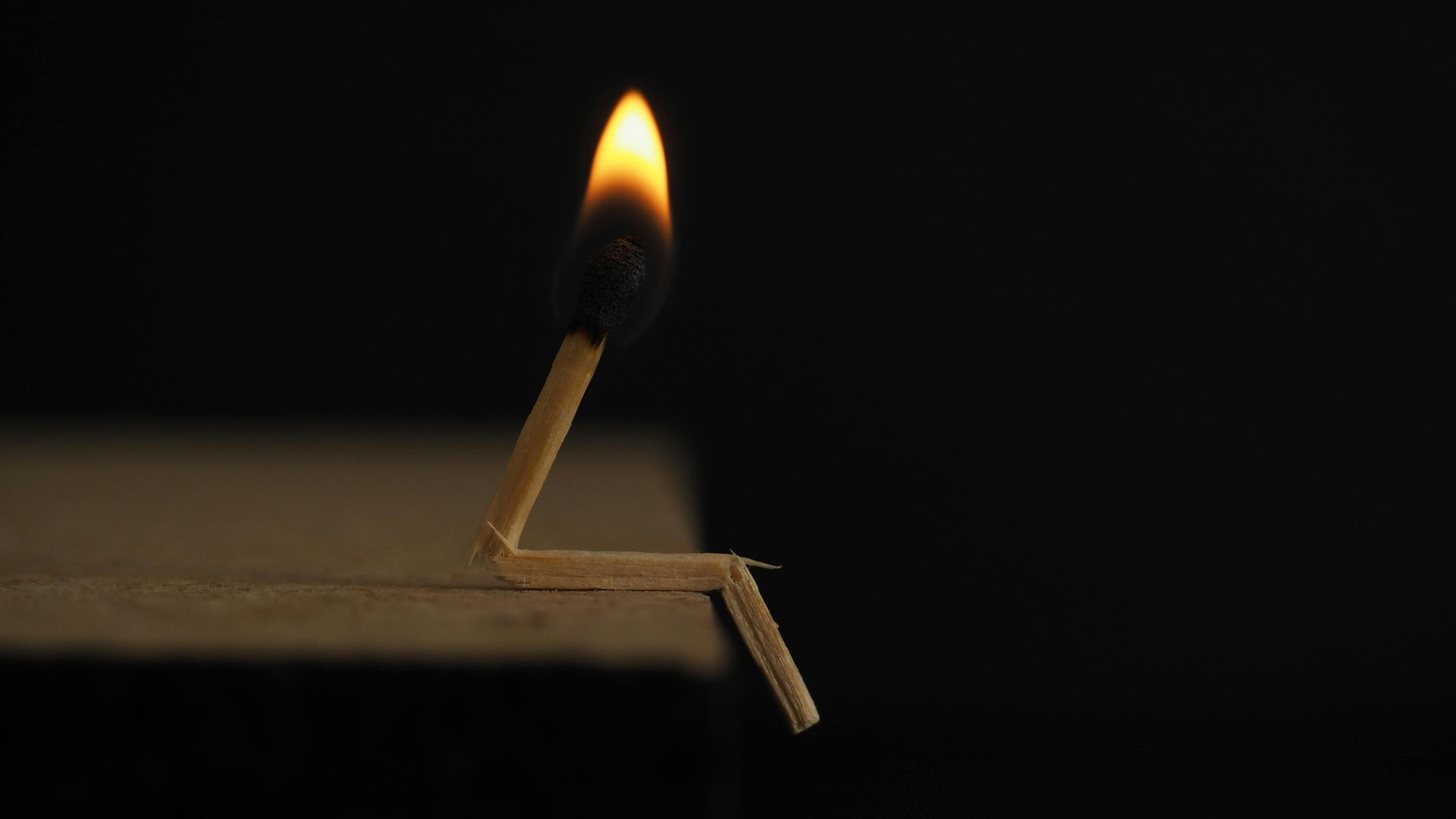 Burning matchstick in the shape of a person, sitting on a ledge against a black background.