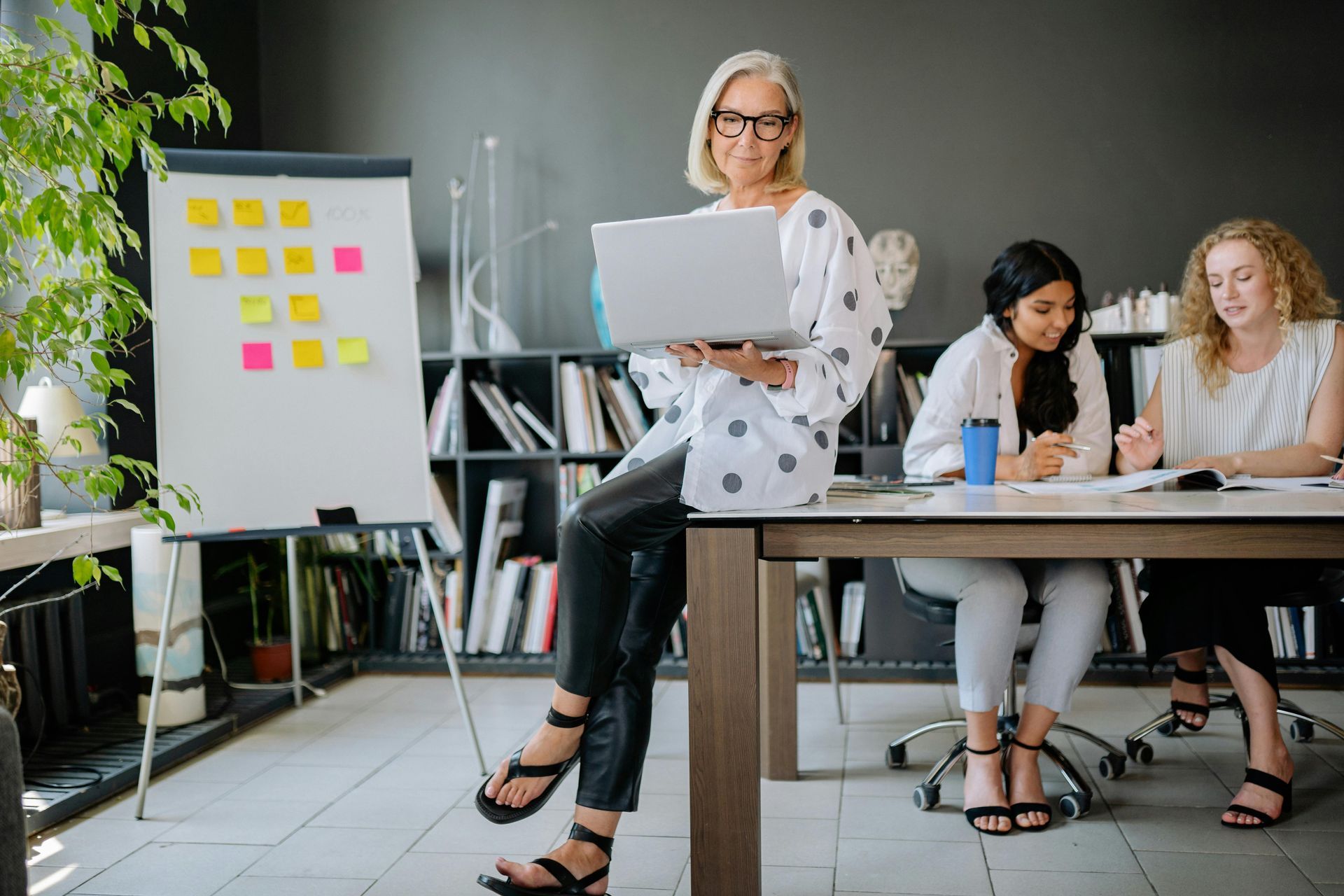 Woman holding laptop, leaning on table in office; two other women working at the table.