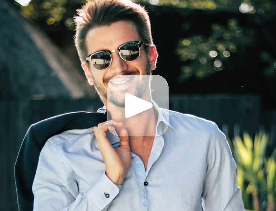 Smiling man in sunglasses, light blue shirt, and jacket over shoulder, outside.
