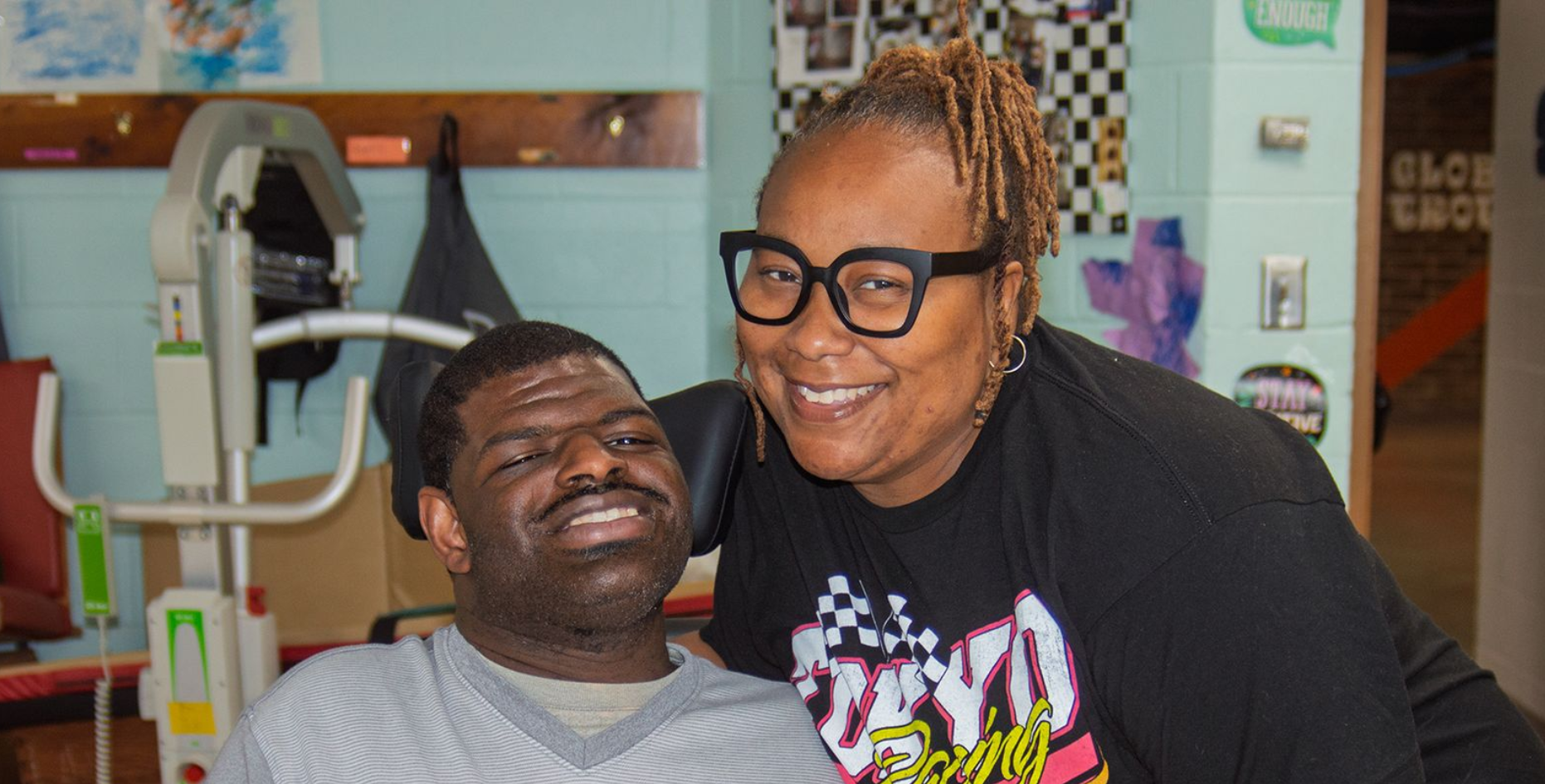 A Direct Support Professional bending down to smile next to a smiling man in a wheelchair.