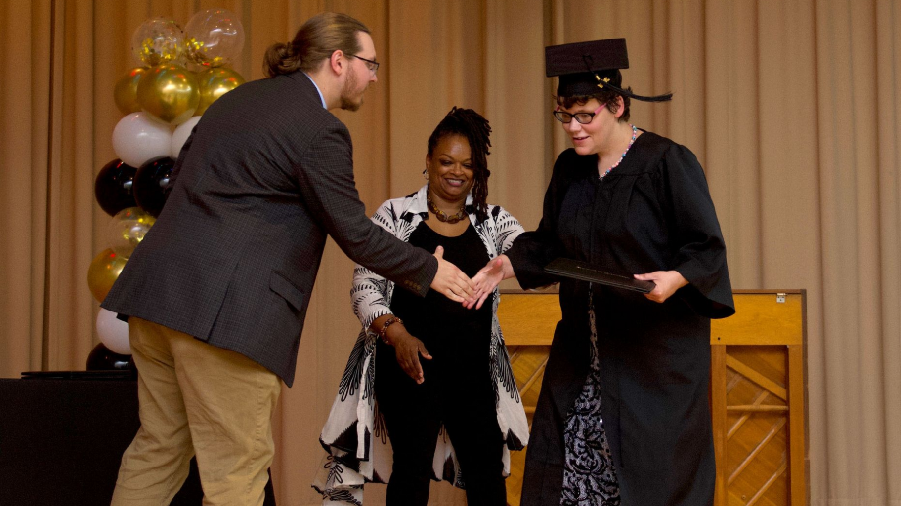 A man in a suit shakes the hand of a woman supported by Orange Grove wearing a cap and gown, as she receives a diploma following the completion of an educational program, as a female staff member smiles in the background.