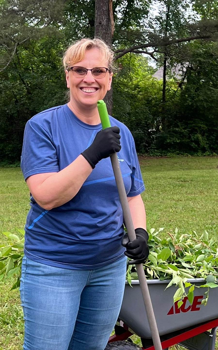 A female volunteer stands smiling, wearing gloves and holding a rake during a landscaping project.
