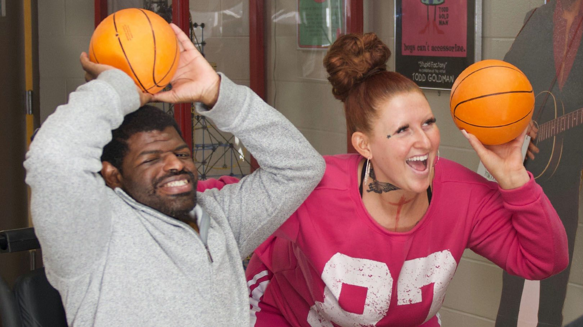 An indivudual served by Orange Grove smiling alongside his direct support staff member as they both hold miniature toy basketballs, preparing to throw them towards a basketball hoop.