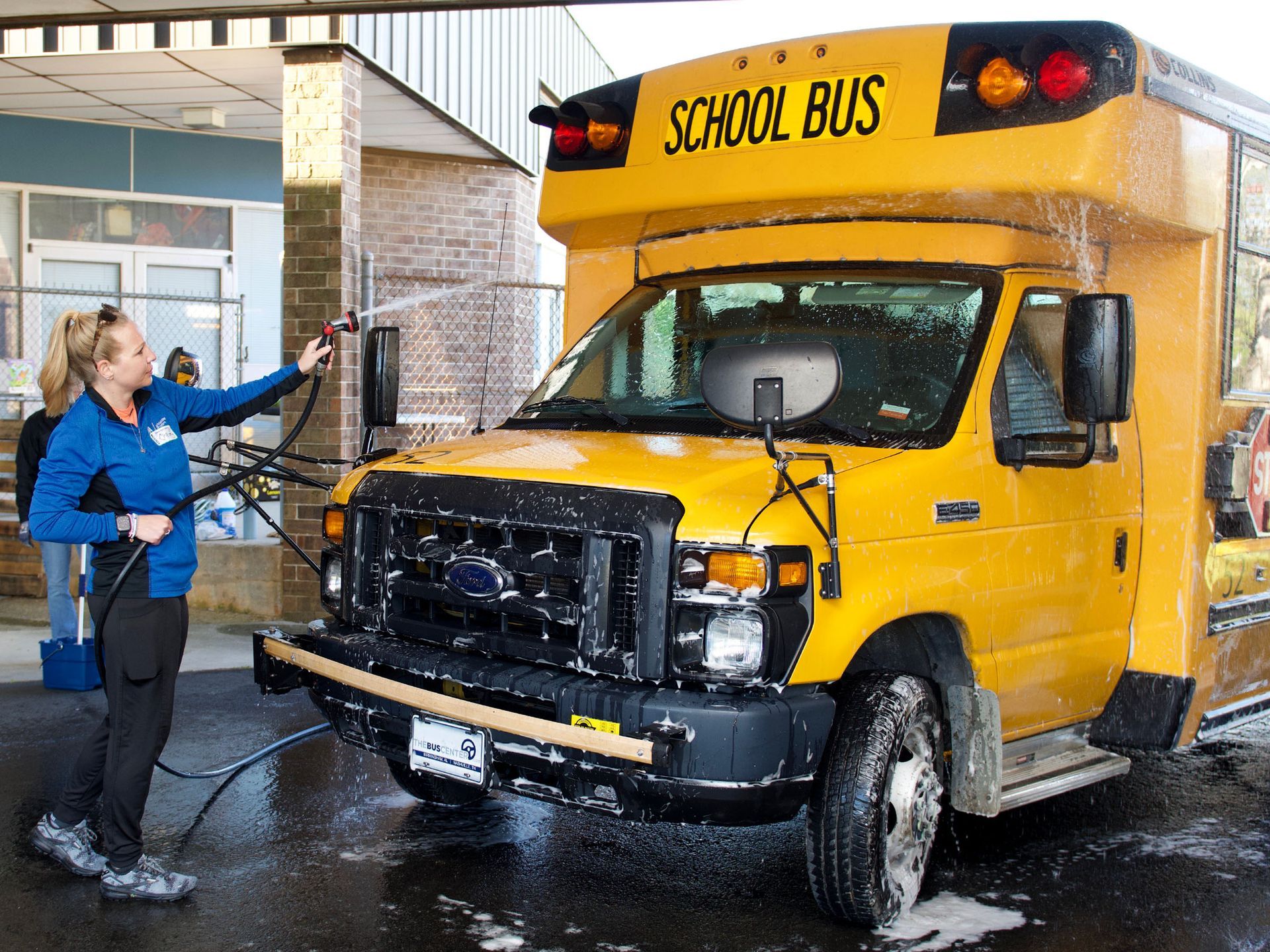 A female volunteer points a water hose at a yellow Orange Grove school bus, washing soap suds off of the front of the vehicle.