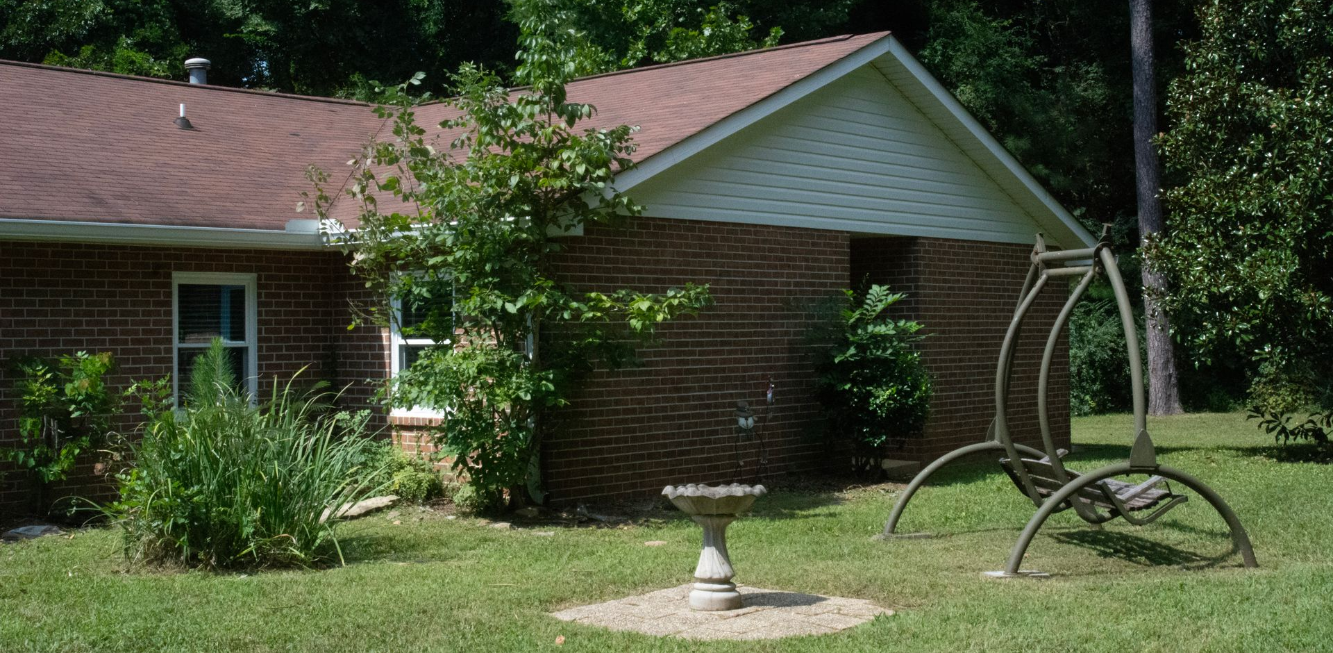 A single-story brick house with white trim, with a grassy yard, a birdbath, and a disability-accessible porch swing.
