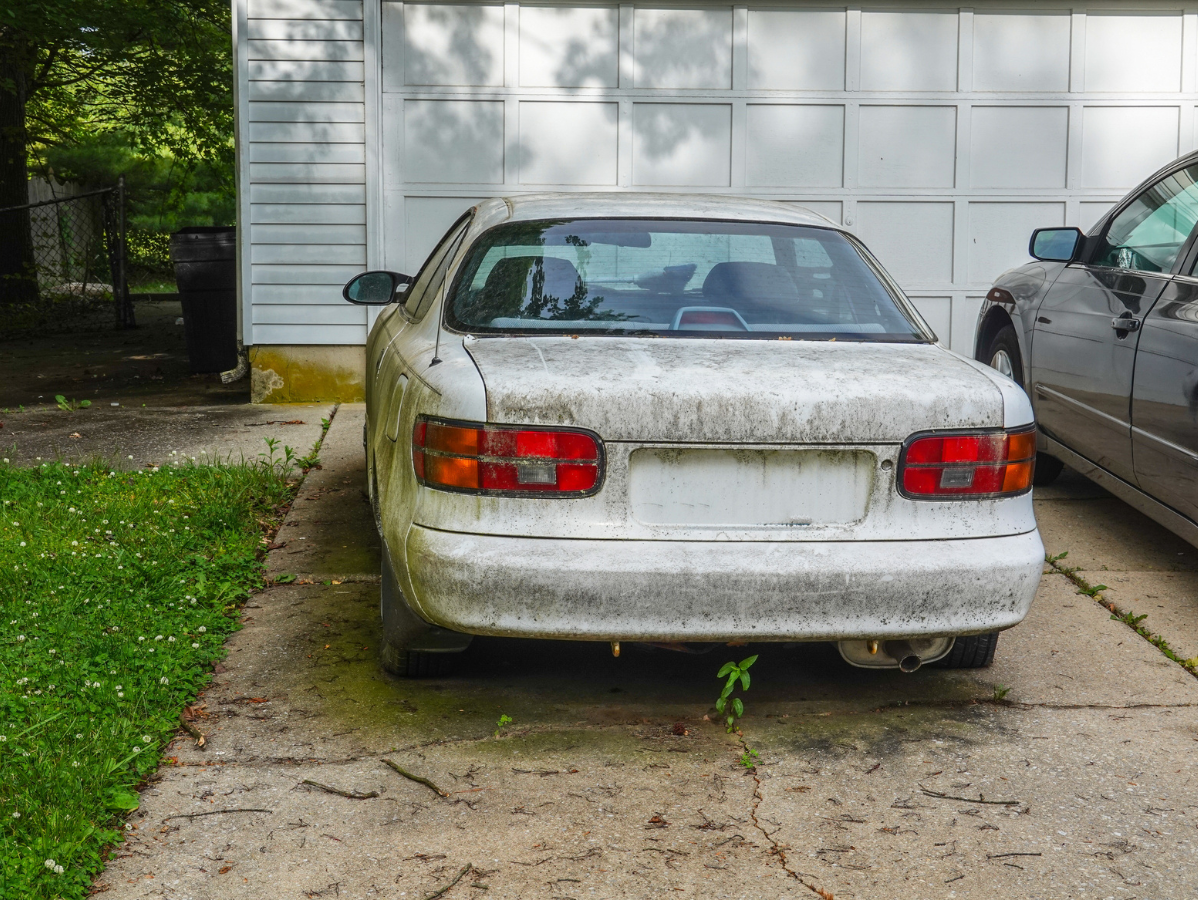 Dusty white sedan parked in a driveway beside a garage and another car