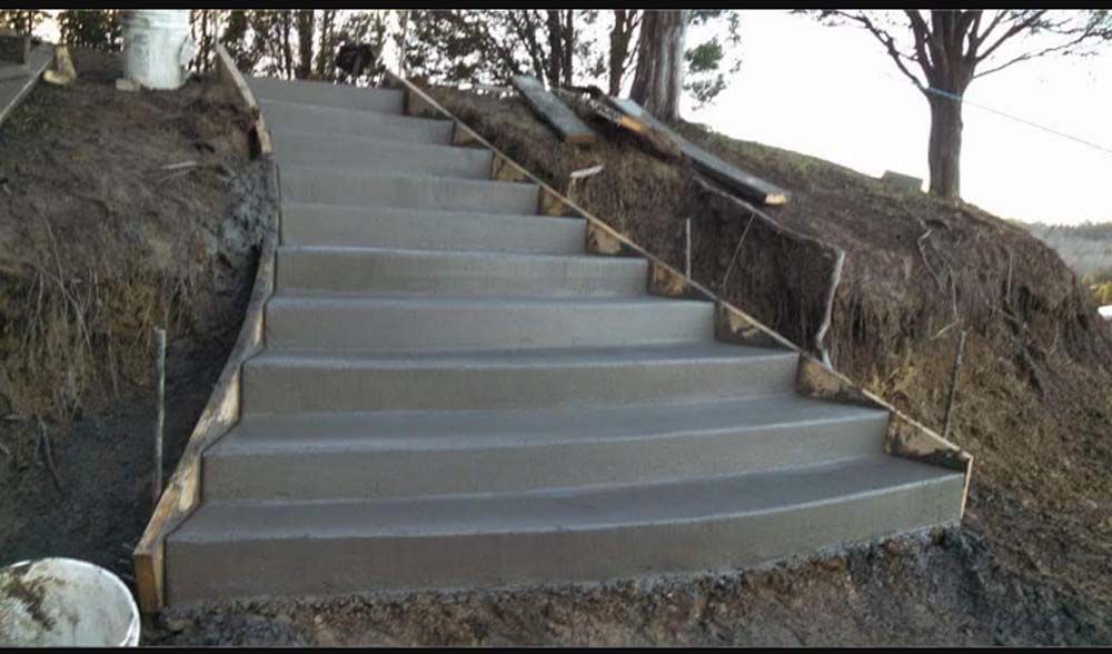 Concrete stairs built into a hillside, bordered by wood forms, surrounded by dirt and trees.