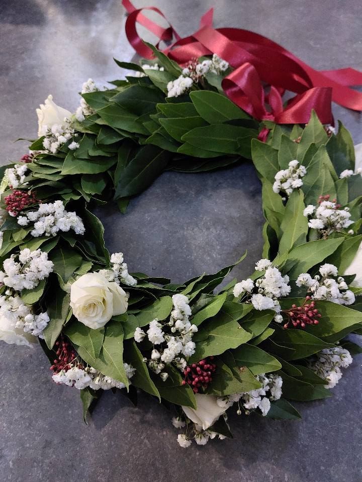 Corona d'alloro con rose bianche, gypsophila e nastro rosso su una superficie grigia.