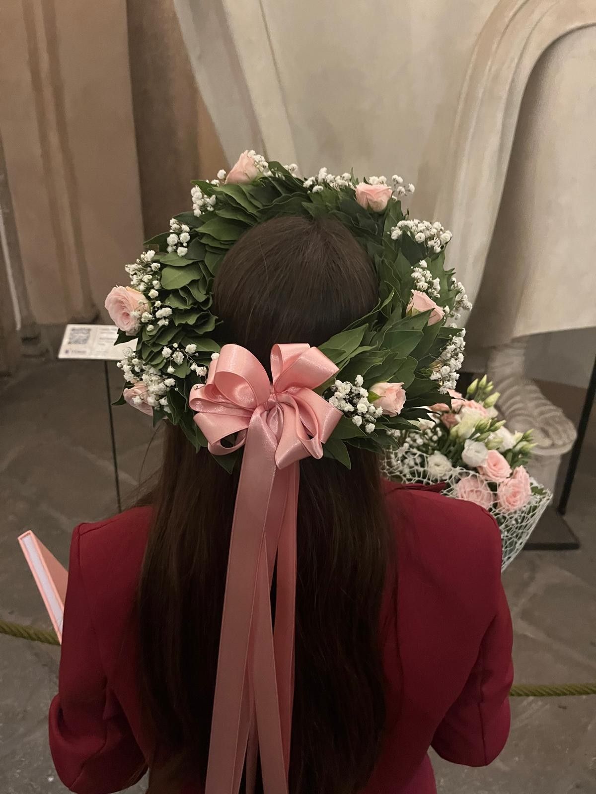 Donna con una corona di fiori e un nastro rosa, vista di schiena, in un ambiente interno. Indossa un cappotto rosso.