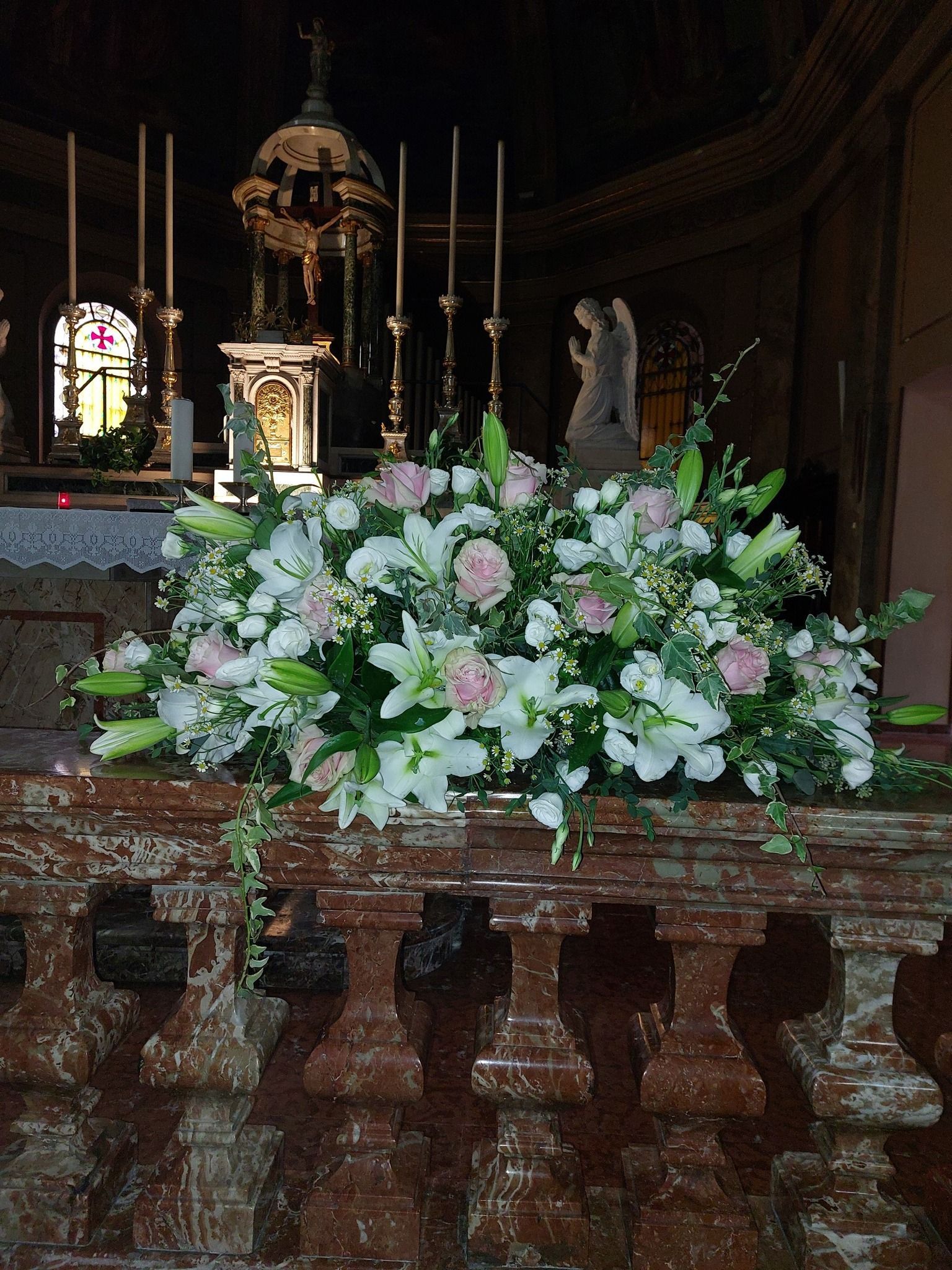 Composizione floreale di gigli e rose sull'altare di una chiesa, con candele e figure religiose sullo sfondo.