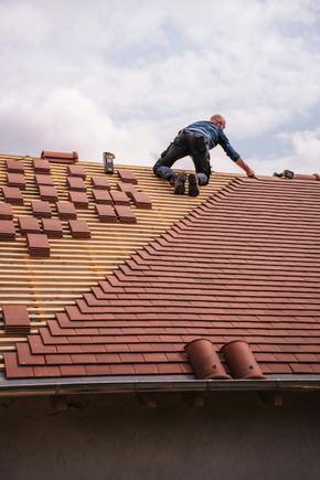 Roofer on a roof, installing red tiles. Partially tiled, cloudy sky.