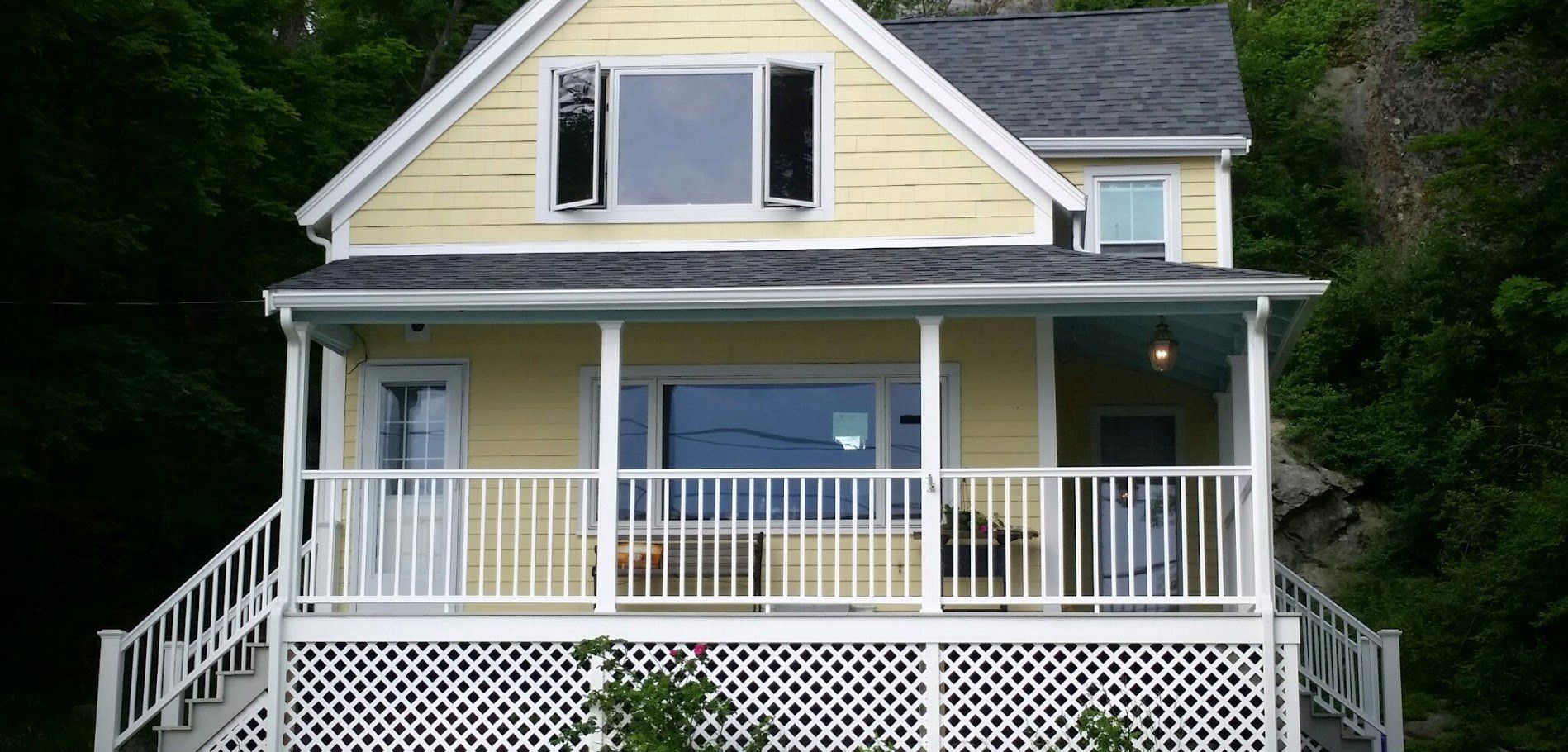 Yellow house with white porch railing and dark shutters on a wooded hillside