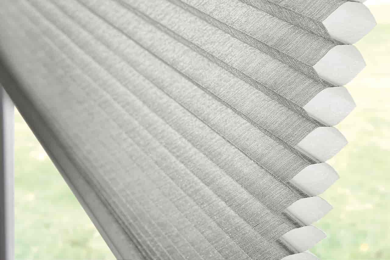Close-up of light gray honeycomb cellular shades, angled against a blurred background. Shades & Awnings | MasterCraft Roofing, Siding & Windows | Serving Eastern MA, NH & Selected Areas of VT