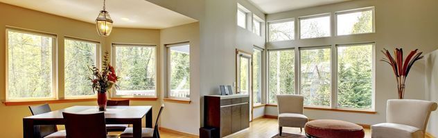 Bright living room with large windows, beige walls, dark dining table, and a view of trees outside