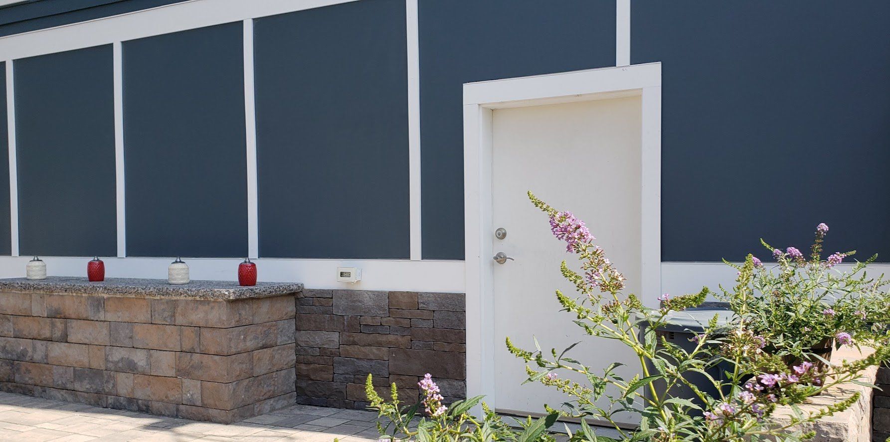 Exterior of a building with blue walls, white trim, and a white door, with a stone planter and flowers in front. Siding | MasterCraft Roofing, Siding & Windows | Serving Eastern MA, NH & Selected Areas of VT