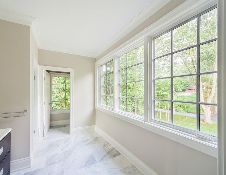 Sunroom with three large windows overlooking a green yard, white trim, beige walls, and marble flooring. MasterCraft Roofing, Siding & Windows | Serving Eastern MA, NH & Selected Areas of VT