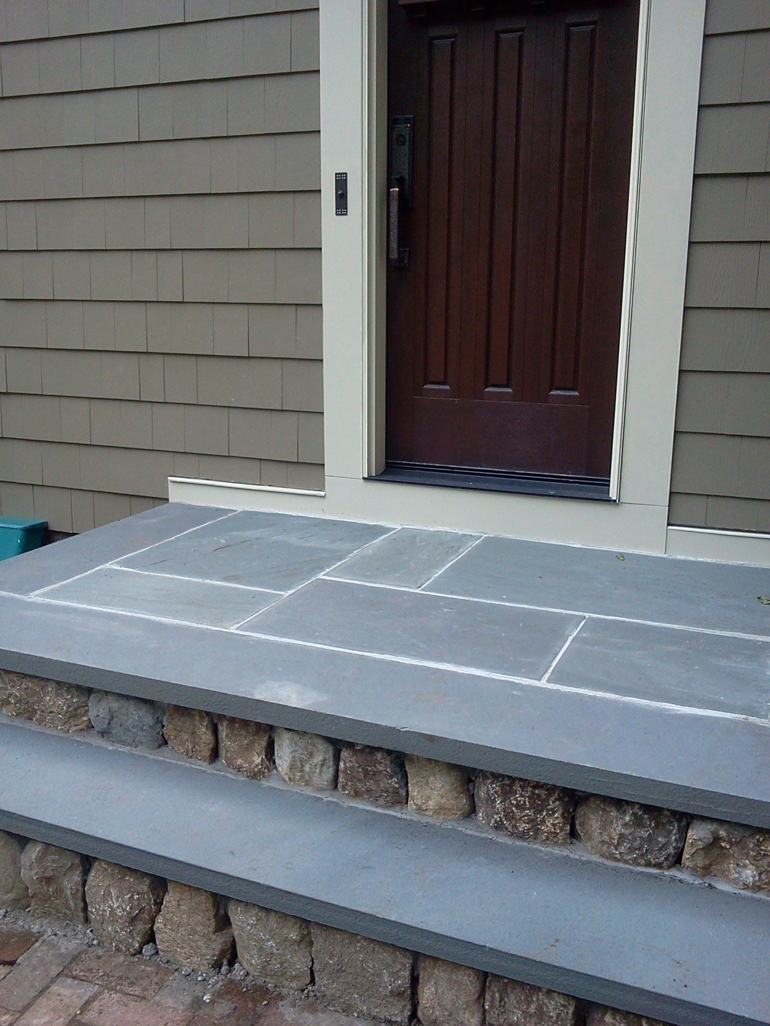 Front porch with gray pavers, stone trim, and a dark front door on a house exterior.
