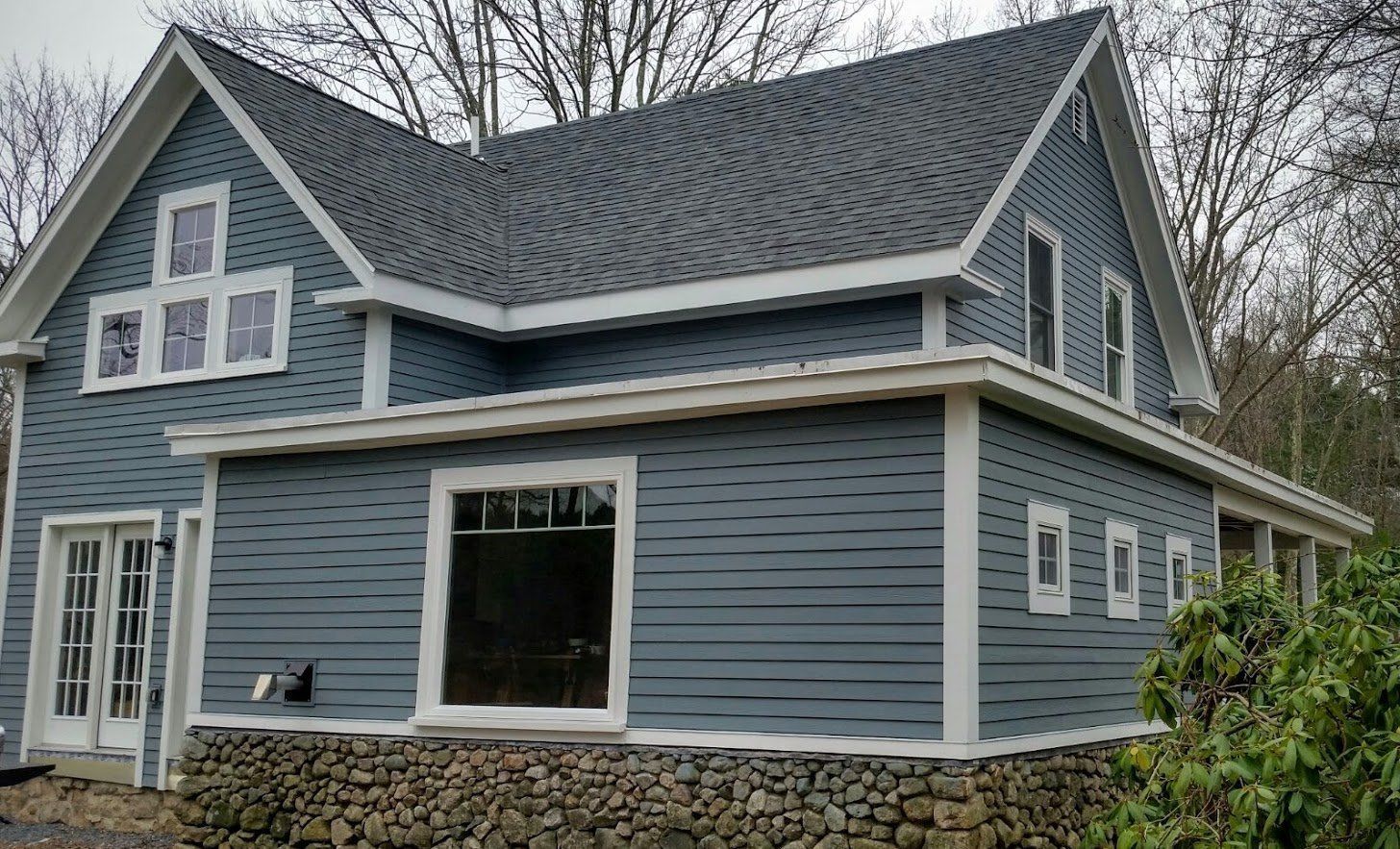 Blue-gray two-story house with white trim, stone foundation, and a large front window in a wooded setting