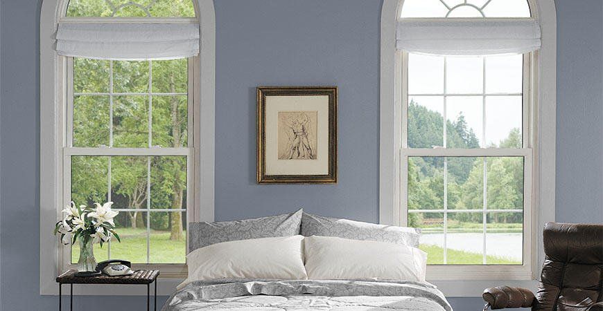 Bright bedroom with a bed between two tall arched windows and blue-gray walls.