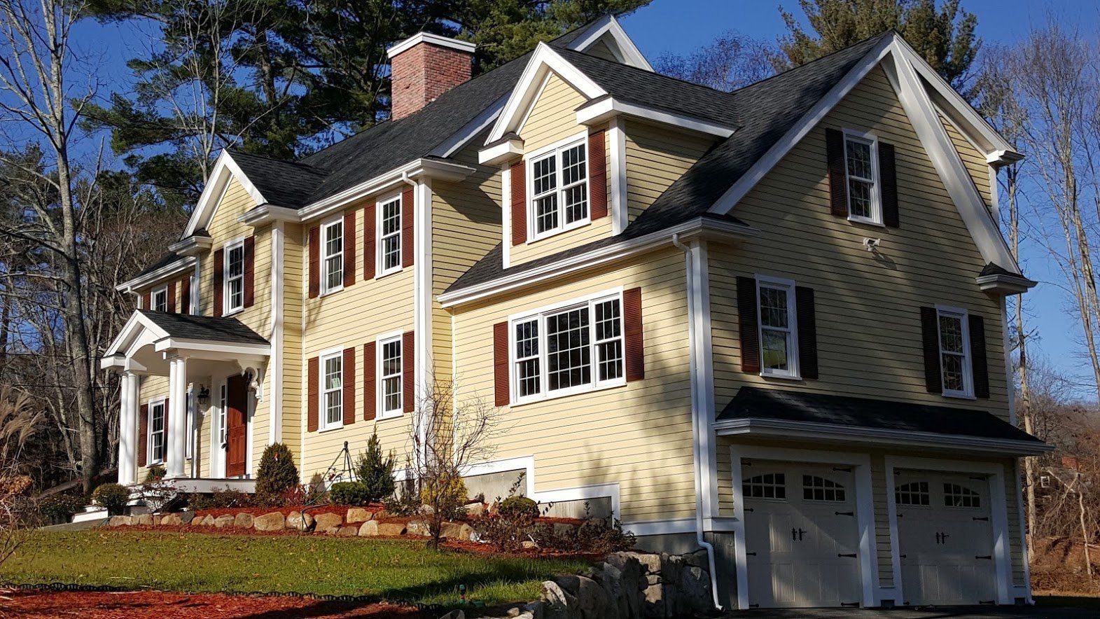 Yellow two-story house with white trim, steep roof, and garage, set among trees in autumn.