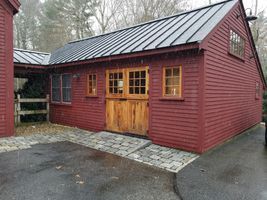 Red wooden shed with double doors and a metal roof beside a wet driveway and stone path