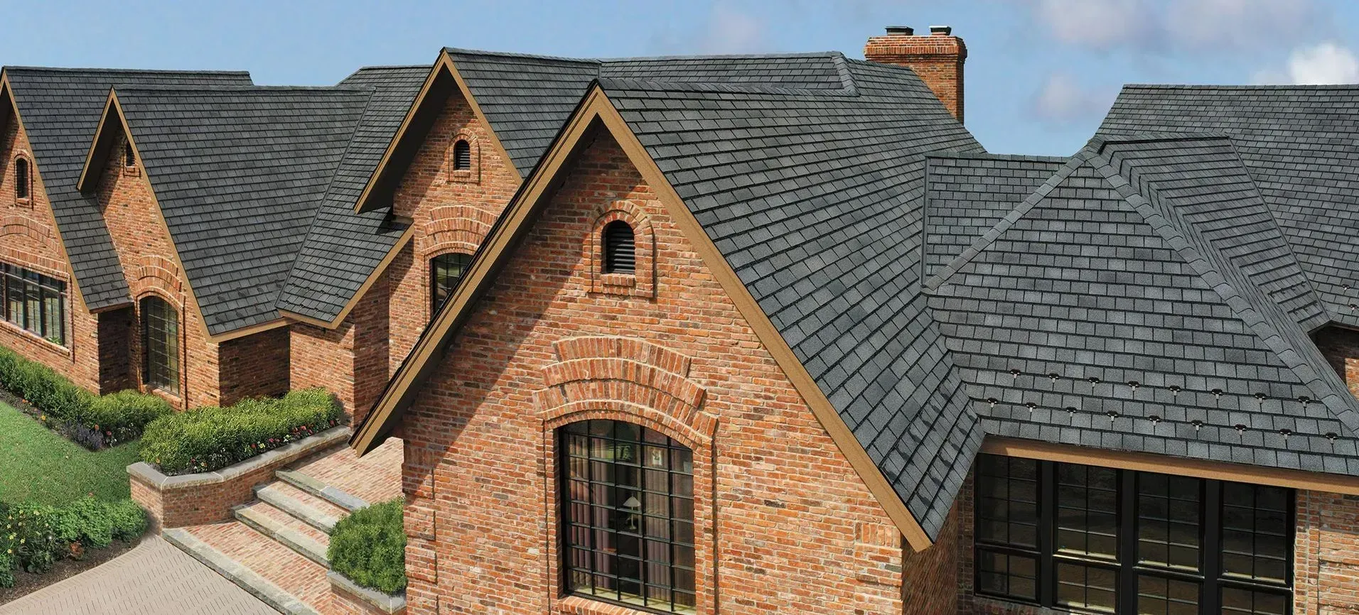 Brick house with dark gray shingle roofs, dormers, and chimney. Roofing | MasterCraft Roofing, Siding & Windows | Serving Eastern MA, NH & Selected Areas of VT