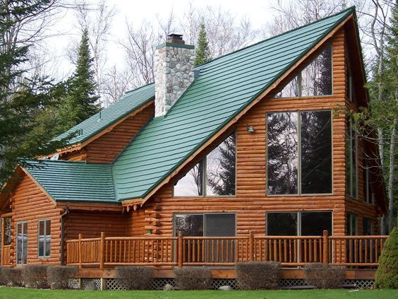 Log cabin with green roof, large windows, and a deck, set in a wooded area. Roofing | MasterCraft Roofing, Siding & Windows | Serving Eastern MA, NH & Selected Areas of VT