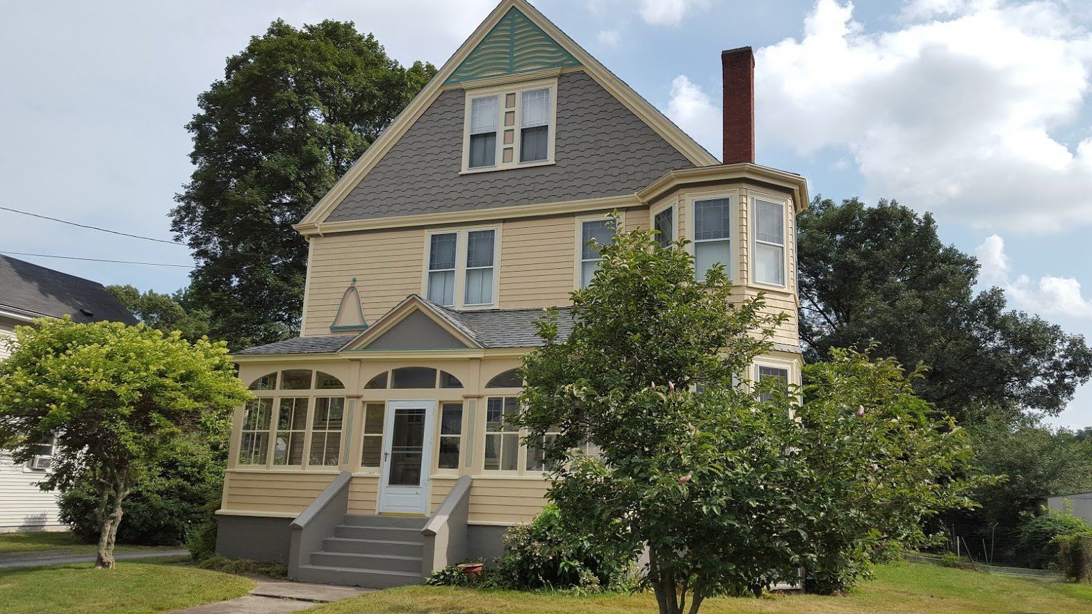 Large gray Victorian-style house with a steep roof, porch, and trees in the front yard